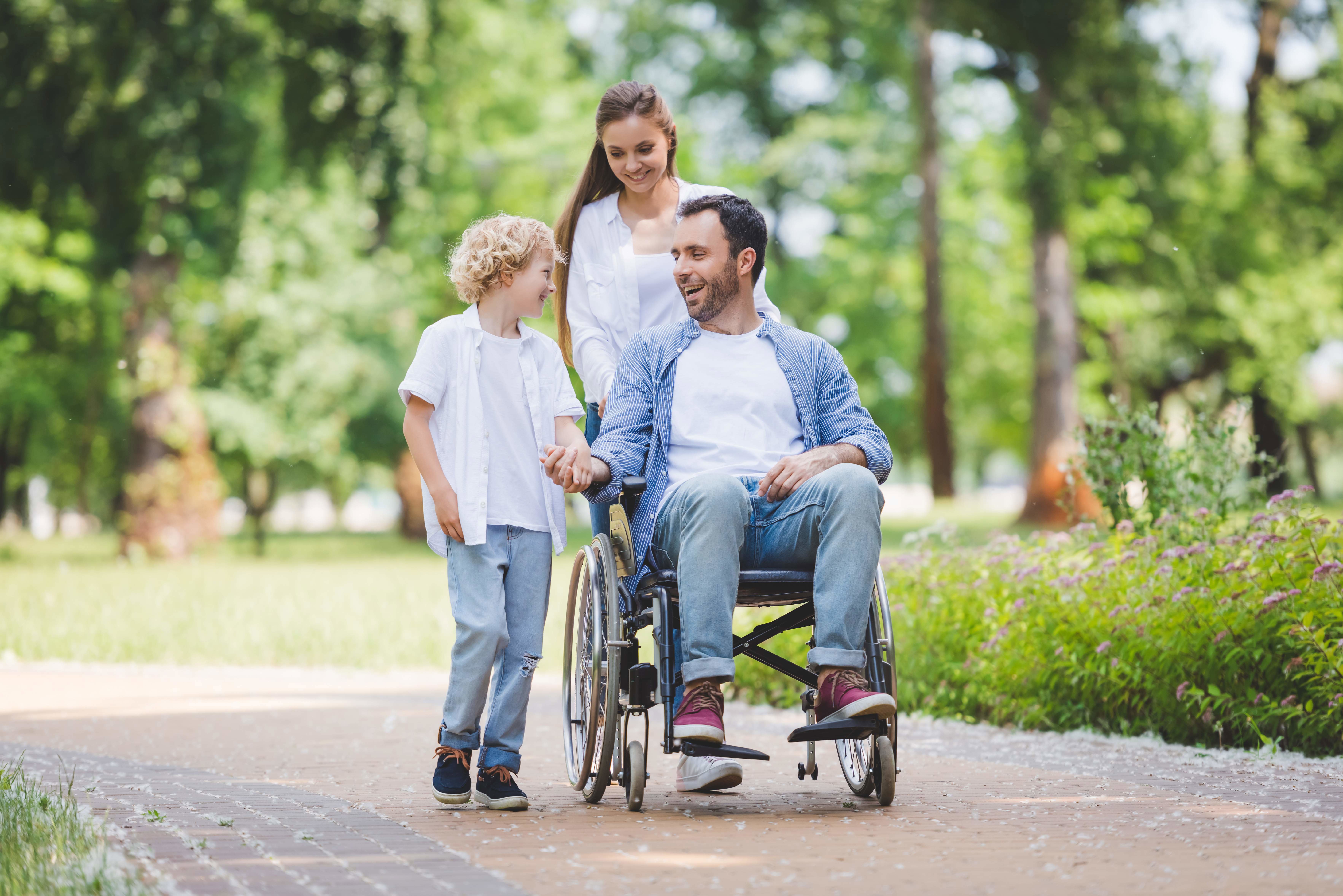 A young family enjoying the outdoors. The father is in a wheelchair, being pushed by his wife while he holds his son's hand